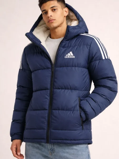 A man wearing an Adidas navy blue hooded puffer jacket featuring white 3-stripes on the shoulders, a white logo on the chest, and a cozy white sherpa fleece-lined hood.