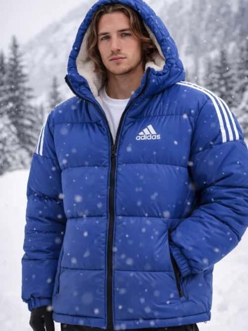 A man wearing a royal blue Adidas hooded puffer jacket with white stripes and a warm sherpa fleece lining in a snowy mountain landscape.