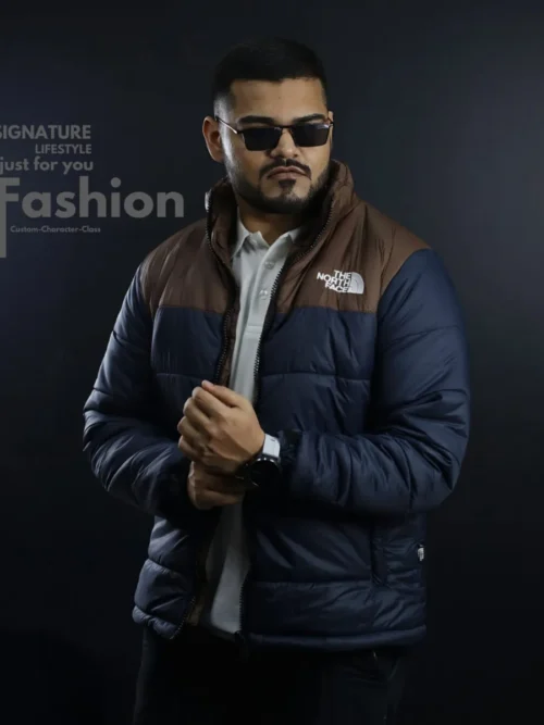 A man wearing a The North Face two-tone navy blue and brown quilted puffer jacket with a white polo shirt and black sunglasses against a dark studio background.