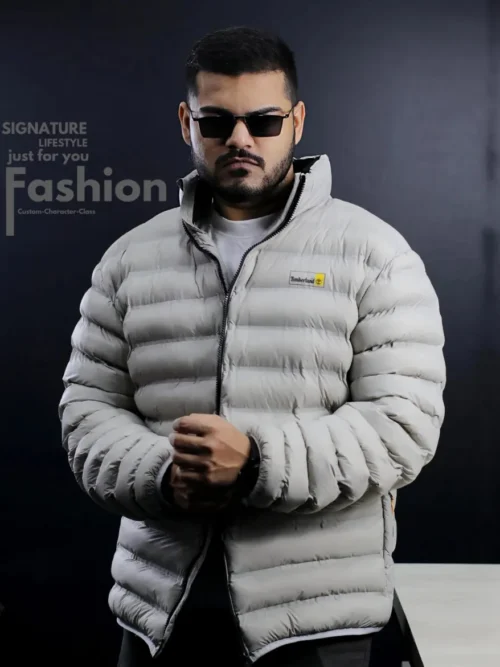A man modeling a premium light grey Timberland puffer jacket with horizontal quilting, black zipper accents, and a stand-up collar.
