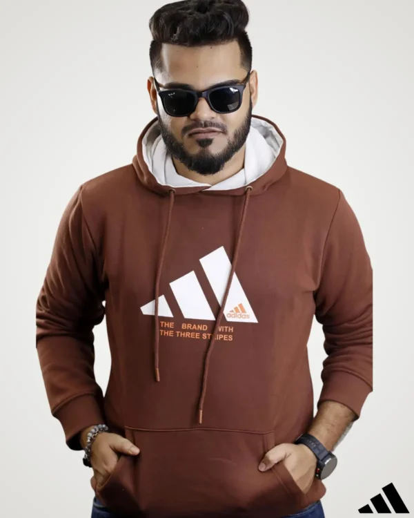 Casual winter outerwear featuring a man in a deep coffee brown pullover hoodie with a white lined contrast hood, front pouch pocket, and bold graphic text print across the front.