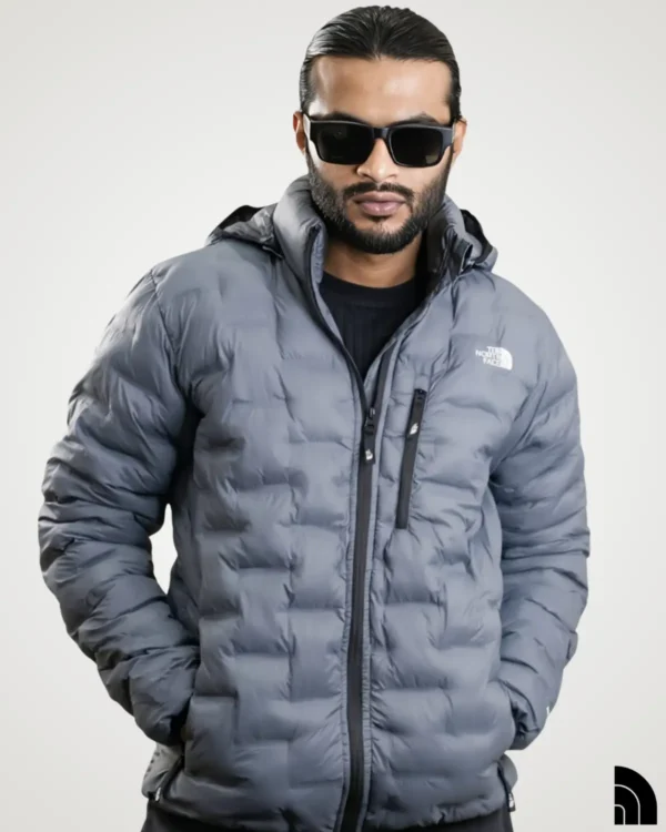 Model with hands in pockets wearing a grey lightweight down jacket for men, designed with horizontal quilted baffles, a protective hood, and dark zipper detailing.