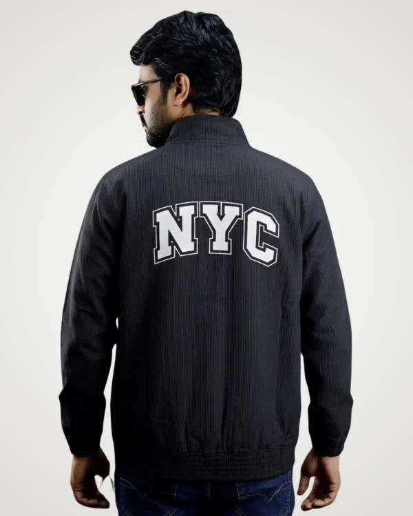 Back view of a model modeling a trendy black corduroy shacket featuring bold white collegiate-style text across the upper back. This fashionable men's corduroy shacket is designed with a protective high neck collar and an elastic hem for a perfect casual fit.