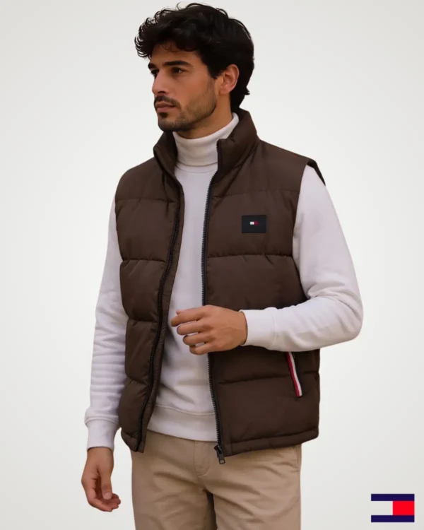 Model posing in a chocolate brown men's puffer vest jacket featuring horizontal padded baffles, high collar neck, zip-up front, and contrast side pocket detailing.