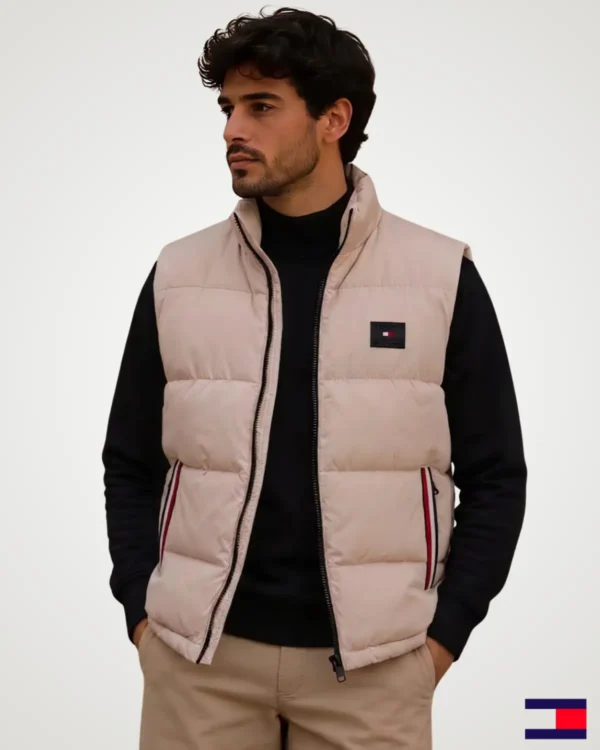Model wearing a light beige-pink men's puffer vest jacket with a stand-up collar, black front zipper, horizontal quilting, and side zippered pockets over a dark inner top.