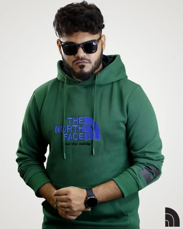 Model posing in a forest green mens typography hoodie, designed with a prominent blue text and geometric logo on the front. This casual drawstring hooded sweatshirt includes a kangaroo pocket and a black square logo patch on the sleeve cuff for a signature look.