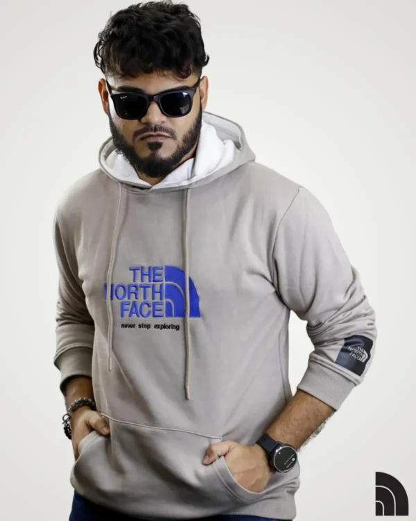 Young man wearing a light grey-beige mens typography hoodie, highlighted by a large blue graphic print that reads 'THE NORTH FACE' and 'never stop exploring'. Features include an adjustable hood, a spacious front pocket, and a black accent patch on the sleeve.