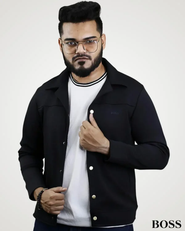 Model holding the lapel of a dark black suede leather jacket for men, designed with a clean spread collar, contrast gold-tone snap buttons, and structured chest yoke.